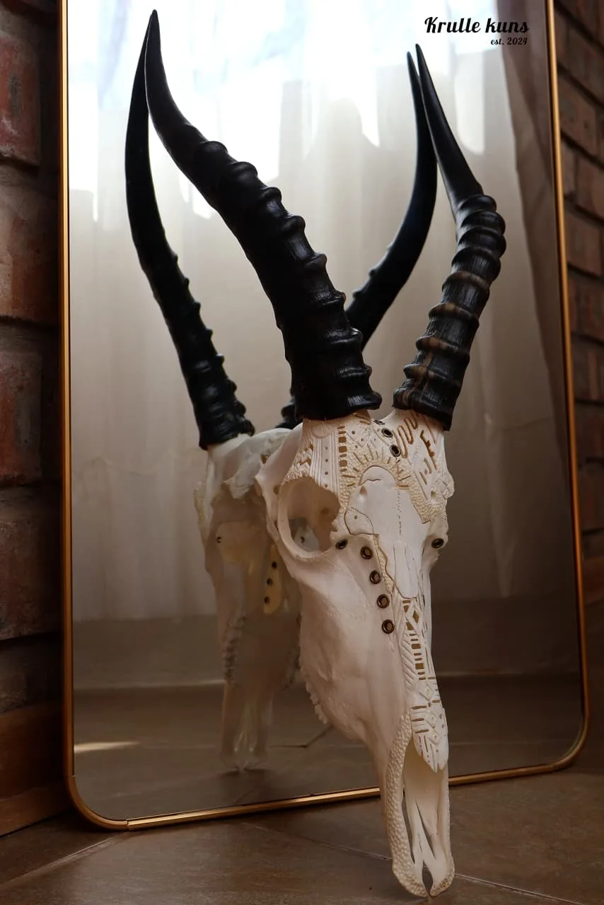 Blesbuck skull with fine carved patterns and brass studs, with long ringed black horns, beside a gold-framed mirror